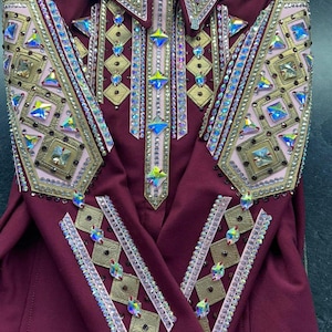 May include: A burgundy equestrian show shirt with elaborate embellishments. The shirt features a collared neckline and is adorned with gold, pink, and iridescent rhinestones in geometric patterns. The sleeves and bodice are heavily decorated.