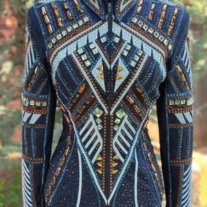May include: A navy blue equestrian show jacket with intricate beadwork and jewel embellishments. The jacket features a high collar, a zipper closure, and geometric patterns in shades of blue, gold, and silver. The sleeves and body are adorned with rows of sparkling accents.