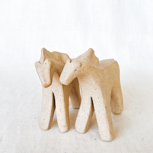 May include: Two ceramic horse figurines in a light beige color with dark speckles. The horses have a minimalist design with elongated legs and a simple, stylized form. The background is a plain white.