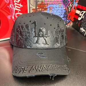 May include: A dark gray baseball cap with the letters "LA" and a skull design. The cap has a distressed brim and the text "FADE AWAKENS" embroidered on the front. Black rhinestones are scattered across the top.