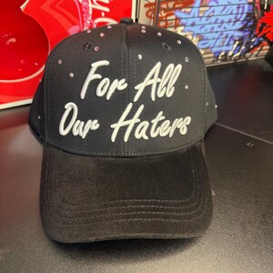 May include: Black baseball cap with the text "For All Our Haters" in white script. The cap is embellished with small, pink rhinestones. The brim is a solid black color.
