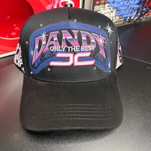 May include: Black baseball cap with the word "DANDY" in pink and blue, accented with rhinestones. The text "ONLY THE BEST" is below the word "DANDY", and the number "52" is at the bottom. The brim is black.