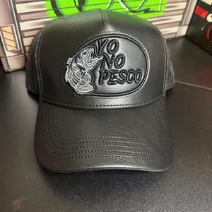 May include: Black baseball cap with a curved brim and mesh back. The cap features an oval patch with the words "YO NO PESCO" and a fish graphic in white stitching.