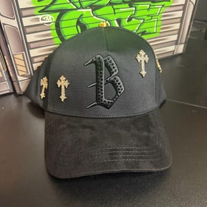 May include: Black baseball cap with a suede brim and a satin crown. The cap features a large, embellished letter "B" and several gold-colored cross pins. The background includes green graffiti art.