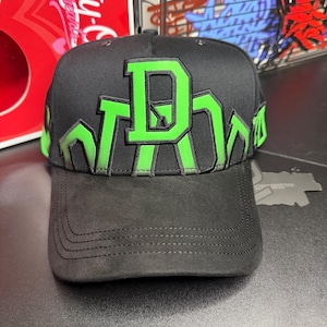 May include: Black baseball cap with the word "DREAM" in bold, neon green lettering across the front. The cap has a flat brim and a structured crown. The design is a mix of street style and bold graphics.