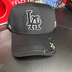 May include: Black baseball cap featuring the letters "LA" in a star-studded design. The cap has a curved brim and embroidered stars. The cap is a dark color.