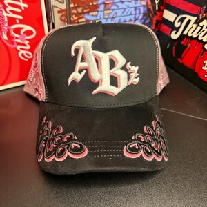 May include: Black and pink baseball cap with the letters "ABz" embroidered in white with pink trim. The brim features pink floral embroidery, and the cap has a black suede-like texture.