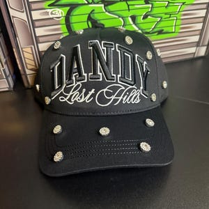 May include: Black baseball cap with the word "DANDY" embroidered in white, along with "Lost Hills" in cursive. The cap is adorned with multiple silver rhinestones. The background features graffiti art.