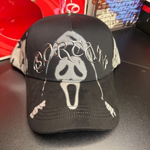May include: Black baseball cap featuring a Scream ghost face design. The word "SCREAM" is in silver lettering across the front. The cap has a black brim and a gray and black patterned mesh back.