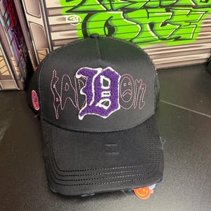 May include: Black trucker hat with a distressed brim. The hat features the word "SAD ON" embroidered in pink and purple thread. A purple "D" is the focal point of the design. The hat has a mesh back.