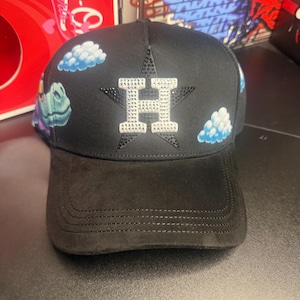 May include: Black baseball cap featuring a rhinestone-studded star and the letter "H". The cap has a curved brim and is decorated with embroidered clouds. The cap is a stylish accessory.