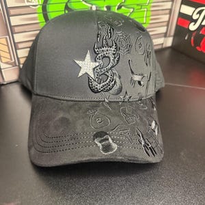 May include: A black baseball cap with a curved brim. The cap features embroidered designs including a star, an octopus, and other symbols. The cap is made of a soft, textured material.