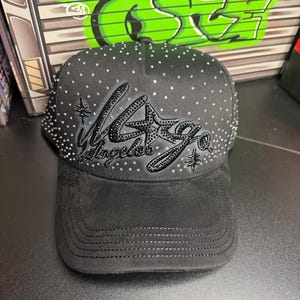 May include: Black baseball cap with the word "Los Angeles" in black rhinestones. The cap has a curved brim and is decorated with small, scattered rhinestones. The hat is made of a suede-like material.