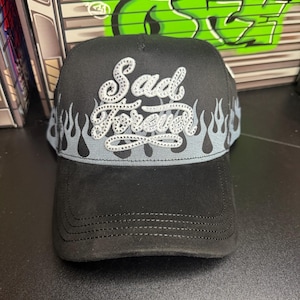 May include: Black baseball cap with the text "Sad Forever" in a cursive font, embellished with rhinestones. The cap features a gray flame design along the brim and is made of a textured fabric.