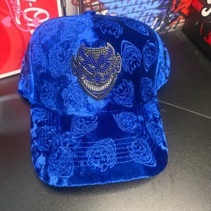 May include: A royal blue baseball cap with a velvet texture. The cap features a repeating pattern and a silver emblem with a blue design on the front. The cap has a curved brim.