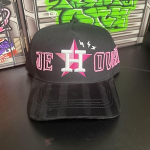 May include: Black baseball cap with a pink star and the letters "JE HOU" embroidered on the front. The brim is black with vertical stripes. The background features graffiti art.