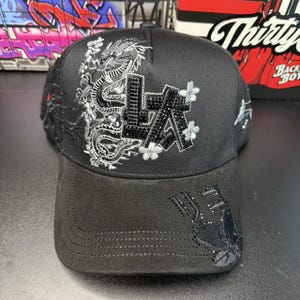 May include: A black baseball cap featuring a silver embroidered dragon design on the left side and the letters LA in black rhinestones. The brim is made of a black suede-like material with additional black rhinestone embellishments on the right side.