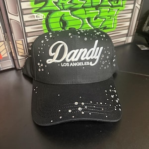 May include: Black baseball cap with the word "Dandy" in white script and "LOS ANGELES" in smaller text. The cap is embellished with small, sparkling rhinestones. The background features green graffiti art.