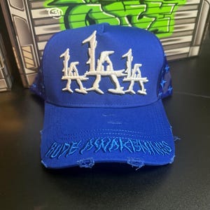 May include: A royal blue baseball cap with white embroidered "LA" lettering on the front. The brim features distressed edges with the embroidered text "HYPE AWAKENS". The cap has a curved brim and a star pattern on the sides.