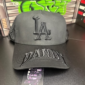 May include: A black baseball cap with the letters "LA" embroidered on the front in a raised, textured design. The brim features the name "DADDY" in a similar style. A tag with a skull and crossbones design is attached.