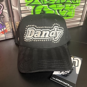 May include: Black baseball cap with the word "Dandy" in silver, outlined with small silver studs. The brim is a darker black suede-like material. A tag with the website www.DANDYHATS.shop is attached.