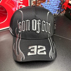 May include: Black baseball cap with the text "SON OF GOD" in silver, outlined with black rhinestones. Silver flame designs are on the sides and the number "32" is on the front. The cap is made of a durable fabric.