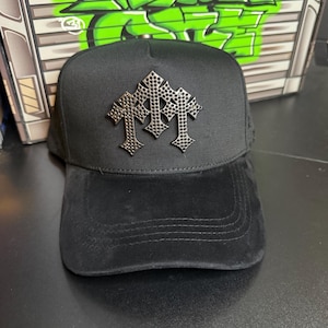 May include: Black baseball cap with a silver cross design. The cap has a curved brim and a structured crown. The background features green graffiti art on a metal surface.