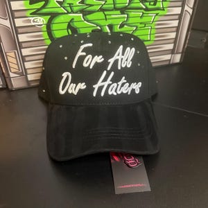 May include: Black baseball cap with the text "For All Our Haters" in white script. The cap is embellished with small, sparkling rhinestones. A tag with a logo and website address is attached.