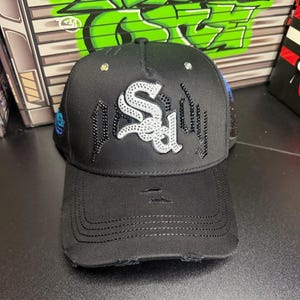 May include: Black baseball cap with a distressed brim. The cap features a white and silver logo with black and silver embellishments. The top of the cap has small, clear rhinestones. The cap is a stylish accessory.