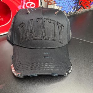 May include: Black baseball cap with the word "DANDY" embroidered across the front. The cap features silver-colored spikes on top and distressed detailing on the brim. The brim has metal accents.