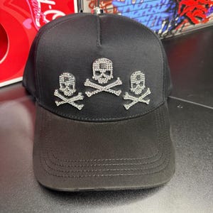 May include: Black baseball cap with three rhinestone skull and crossbones designs. The cap has a curved brim and a structured crown. The skulls are detailed with sparkling rhinestones, adding a touch of glamour to the edgy design.