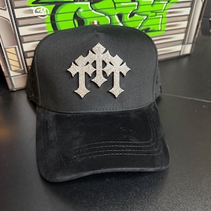 Puede incluir: Gorra de béisbol negra con visera de terciopelo y corona estructurada. La gorra presenta un emblema plateado de tres cruces incrustadas con pedrería. Un accesorio elegante.