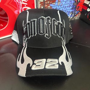 May include: Black baseball cap with the text "SON OF GOD" in white, gothic-style lettering. The brim features a flame design and the number "92" in white. The cap has a flat brim and a structured crown.