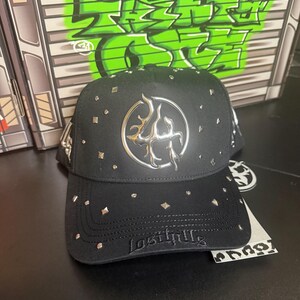 May include: Black baseball cap with silver studs and a silver emblem on the front. The brim has the embroidered text "Last Pills". The background features green graffiti-style text on a gray surface.