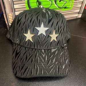 May include: Black baseball cap with a textured pattern and three star embellishments. The stars are gold and silver. The cap has a curved brim and a metal clasp.