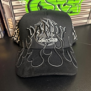 May include: Black baseball cap with the word "Dandy" in a stylized font, with "Lost Hills" underneath. The cap features flame designs outlined in white stitching. The background includes graffiti art.