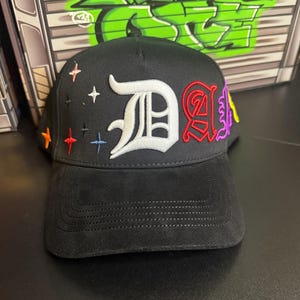 May include: Black baseball cap with a suede brim. The cap features a white embroidered "D" and a red and purple embroidered "A". Colorful star and cross accents are embroidered on the cap. The cap is a stylish accessory.