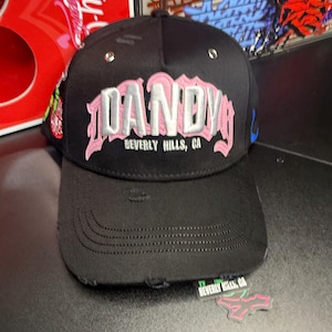 May include: Black baseball cap with the word "DANDY" in white and pink embroidery. The text "BEVERLY HILLS, CA" is embroidered below. The cap has a distressed brim and a small tag that reads "BEVERLY HILLS, CA".