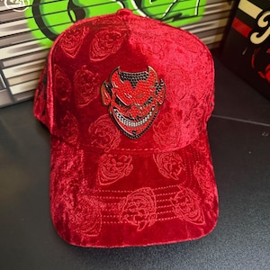 May include: A red velvet baseball cap with a devil-themed emblem. The cap features an embossed pattern and a black and red devil pin with rhinestone accents. The hat has a curved brim and a structured crown.