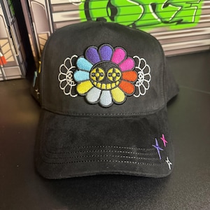 May include: Black baseball cap with a colorful flower design embroidered on the front. The flower has petals in various colors, including blue, yellow, and red. The cap also features embroidered crosses in pink, purple, and white.