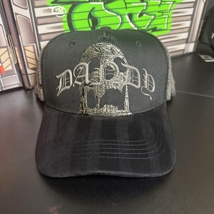 May include: Black trucker hat with a silver embroidered design featuring the word "DADDY" in gothic lettering. The hat has a black brim and a mesh back. The design includes a skull and architectural elements.