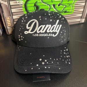 May include: Black baseball cap with the word "Dandy" in white script and "Los Angeles" below. The cap is embellished with scattered rhinestones. The brim is curved and the cap is sitting on a black surface.