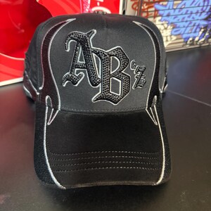 May include: Black baseball cap with the letters "ABz" in a decorative font, embellished with black rhinestones. The cap features a curved brim and white stitching accents along the edges, with a flame-like design on the sides.