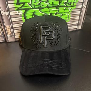 May include: Black baseball cap with a suede brim and a structured crown. The cap features a large, embellished "PF" logo in the center, surrounded by small, sparkling accents. The background includes green graffiti art.