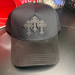 May include: Black baseball cap with a suede brim and a mesh back. The cap features a silver emblem of three crosses. The cap has a curved brim and a snap closure at the back.