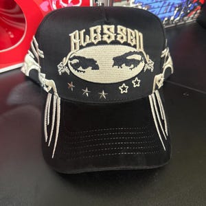 May include: Black baseball cap with the word "BLESSED" embroidered in beige. The cap features a design with two animal silhouettes and stars. White decorative stitching accents the sides and brim.