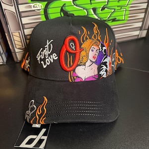 May include: Black baseball cap with embroidered design. Features the text "First Love" and a graphic of a woman with flames. The cap has a curved brim and a tag attached.