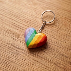 Può includere: Un portachiavi a forma di cuore con una finitura liscia e lucida, caratterizzato da strisce diagonali dipinte nei colori dell'arcobaleno, tra cui rosso, arancione, giallo, verde, blu e viola. Il cuore è fissato a un anello metallico argentato.