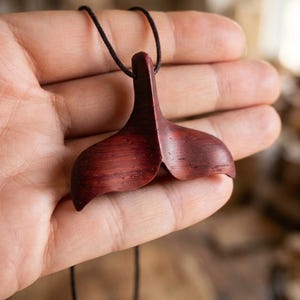 Pode incluir: Um pingente de madeira castanho-avermelhado escuro esculpido no formato de uma cauda de baleia, suspenso por um cordão preto fino. O veio da madeira é visível na superfície lisa e curva do pingente, que apresenta um entalhe vertical.