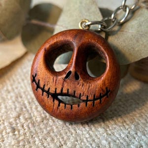 May include: A round, wooden pendant carved with a smiling face design. The pendant is brown with a stitched mouth and eye sockets. It is attached to a silver chain. The design is reminiscent of a character from a popular animated film.